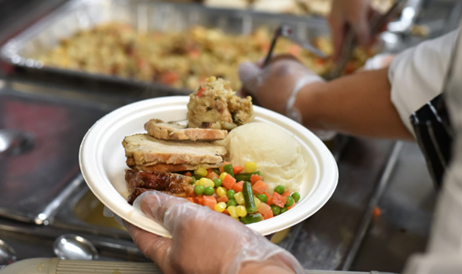 Person serving a plated meal with vegetables, mashed potatoes, and meat at a community meal service.