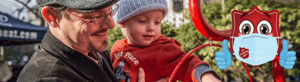 An adult holding a child as the child reaches toward a smiling Salvation Army kettle character wearing a face mask